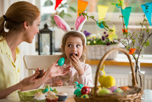 Mother And Daughter Celebrating Easter, Eating Chocolate Eggs. Happy Family Holiday. Cute Little Girl In Bunny Ears Laughing, Smiling And Having Fun.