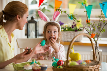 Wall Mural - Mother and daughter celebrating Easter, eating chocolate eggs. Happy family holiday. Cute little girl in bunny ears laughing, smiling and having fun.