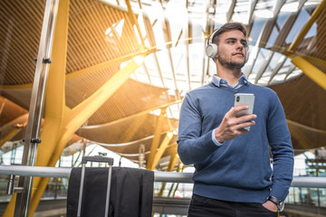 young handsome man happy listening music smiling and using his mobile phone at the airport waiting his flight with luggage