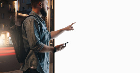 Summer night. Young hipster bearded man stands on city street and touches large glowing touchscreen while holding smartphone. Innovative technologies, digital display, urban navigation. Mock up.