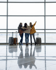 Backs of young companions with suitcases watching aircrafts landing and taking off. One girl is pointing with fingers. Copy space