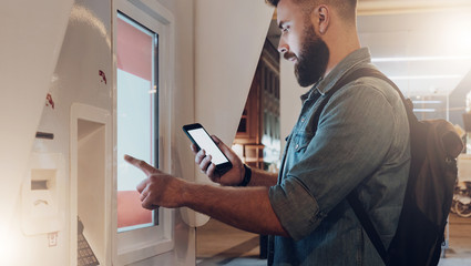 Summer night. Young hipster man with beard stands on city street and touches digital display while looks on screen of smartphone. Innovative technologies, digital display, urban navigation. Mock up.