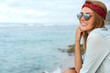 © WHstudio Leushin N - Glad young cute female hiker wears sunglasses and headband, sits on cliff, looks at beautiful view of peaceful ocean or sea with copy space for your text or advertisment. Seascape and nature.