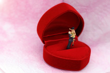 Miniature People: Couple Standing On Red Wedding Ring Box. Concept Of Marriage, Engagement Or Love.