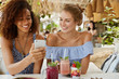 © WHstudio Leushin N - Photo of pleasant looking African American female blogger shows her own web site on cell phone to friend, shares great success and many followers. Happy women watch funny pictures on mobile phone