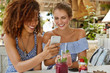 © WHstudio Leushin N - Positive young women recreat together at cozy restaurant, read good news online via cell phone, enjoy tasty cocktails or view funny photos from their excursion. Multiracial relationship concept