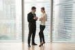 © fizkes - Female and male colleagues discussing work while standing near large window with skyscrapers view outside. Company leader planning daily meetings, giving instructions to secretary with tablet in hands