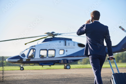 Fotografía Rear View Of Businessman Walking Towards Helicopter Whilst Talking On Mobile Pho