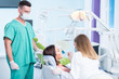 © Senad - people, medicine, stomatology and health care concept - happy female dentist with mirror checking patient girl teeth up at dental clinic office while male assistant is waiting behind.