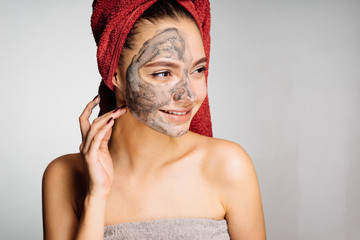 happy young girl with a red towel on her head puts a useful mask on the face, wants to be beautiful, smiles