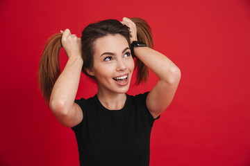 Sticker - Cheerful childish woman 20s in black t-shirt having fun and making two ponytails with her brown hair, isolated over red background