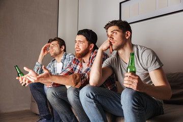 Wall Mural - Photo of discontented adult men expressing frustration, while sitting on sofa and watching football game in apartment