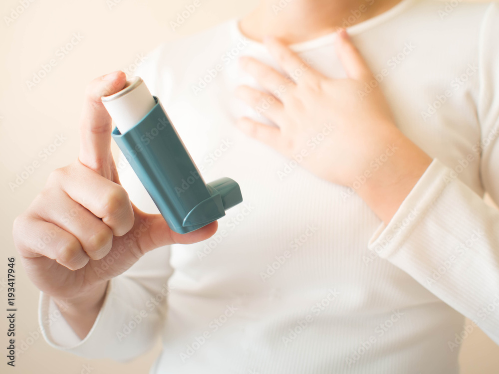 Young female in white t-shirt using blue asthma inhaler for relief asthma attack. Pharmaceutical products is used to prevent and treat wheezing and shortness of breath caused asthma or COPD. Close up. - obrazy, fototapety, plakaty 