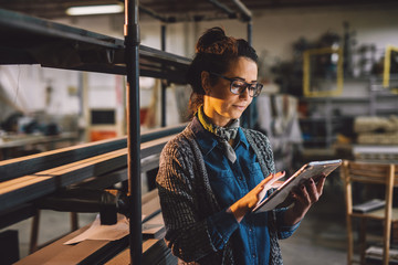 Close up view of hardworking focused professional motivated business woman holding a tablet next to the shelf with metal pipes in the fabric workshop.