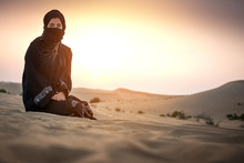 Young Woman Bedouin In Black Traditional Clothes Against Sunset Sky Over Desert.