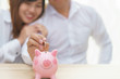 © petzshadow - Smile couple putting a coin into a pink piggy bank on wooden desk - save money for the future.
