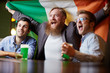 © pressmaster - Three excited fans with national irish flag sitting in pub and watching football match