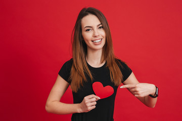 Poster - Romantic woman 20s with long brown hair smiling and gesturing finger at paper heart, isolated over red background