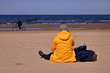 Samotna kobieta siedzi tyłem na piasku na plaży, w żółtej kurtce, obok niej leżą torby, w tle nieostry rowerzysta jedzie brzegiem morza, słonecznie
