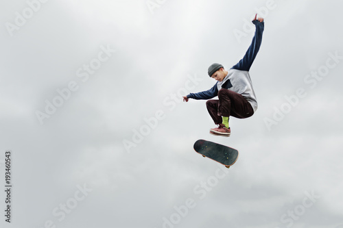 Fotografia A skater teenager in a hat and a sweatshirt does a trick in the air with a flip of the board against a cloudy gray sky