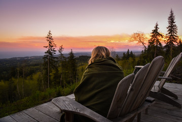 Woman in blanket on wooden chair watches sun rise over forest