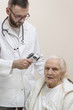 Lekarz w białym fartuchu mierzy temperaturę termometrem laserowym bardzo starej siwej kobiecie. Brodaty lekarz w białym kitlu ze stetoskopem na szyi mierzy temperaturę bardzo starej siwej kobiecie ter