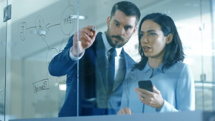 Wall Mural - Top Manager and Female Executive Outlining Company Optimization Plan on a Glass Blackboard, Drawing with Market and Using Smartphone while Standing in the Modern Glass and Concrete Office. 