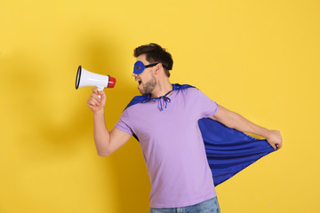 Young man in superhero costume with megaphone on color background