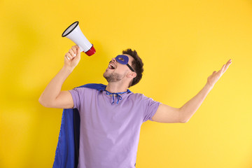 Wall Mural - Young man in superhero costume with megaphone on color background