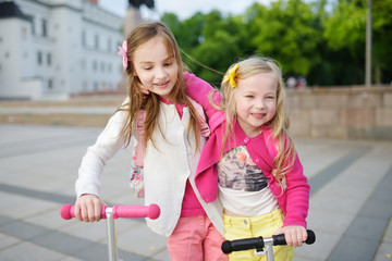 Wall Mural - Small children learning to ride scooters in a city park on sunny summer evening. Cute little girls riding rollers.