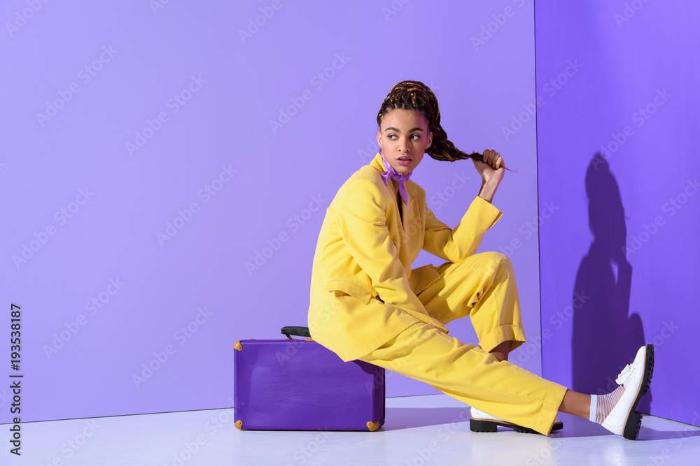 african american girl posing in yellow suit sitting on purple suitcase, on trendy ultra violet background - obrazy, fototapety, plakaty 