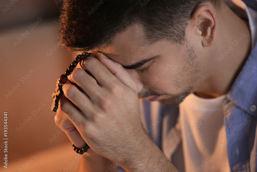 Religious young man with rosary beads praying at home, closeup
