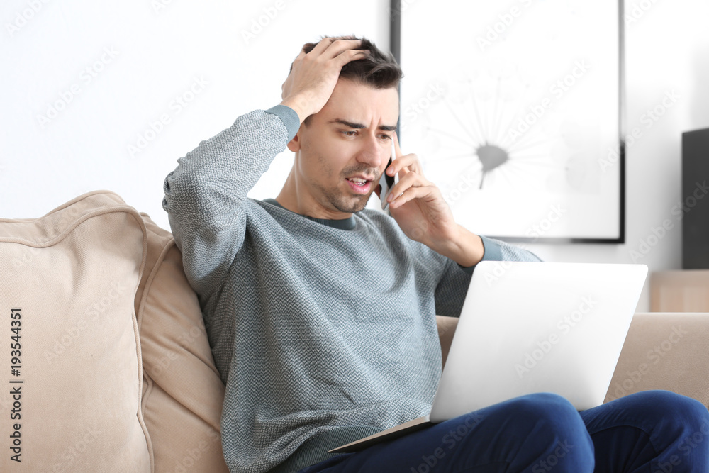 Dissatisfied young man talking on phone while working with laptop at home