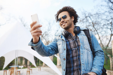 Wall Mural - Outdoor shot of attractive young african-american with afro hairstyle and headphones over neck, wearing trendy clothes and glasses, holding smartphone while recording street band in park.