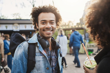 Wall Mural - Positive and charming dark-skinned man with afro hairstyle walking with girlfriend in park, smiling broadly at camera while being in good mood. Siblings spending time together