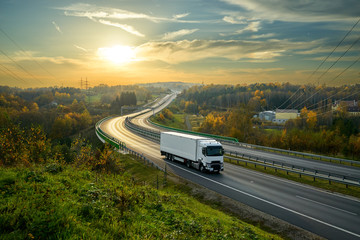 Wall Mural - white truck driving on the highway winding through forested landscape in autumn colors at sunset