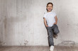 © deniskomarov - Little fashionable boy posing in front of gray concrete wall. Portrait of smiling child in white T-shirt and gray trousers. Concept of style and fashion for children.