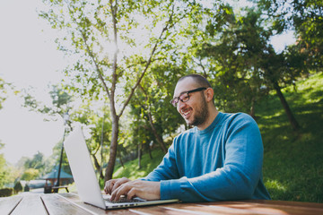 Young successful smart man businessman or student in casual blue shirt glasses sitting at table with mobile phone in city park using laptop working outdoors on green nature. Mobile Office concept.