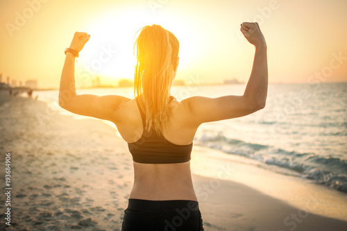 Back view of strong sporty girl showing muscles at the beach during sunset Tablou Canvas