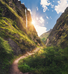 Wall Mural - Amazing Himalayan mountains covered green grass, high waterfall, beautiful path, green trees, blue sky with yellow sun and clouds in Nepal at sunset. Mountain canyon. Travel in Himalayas.Landscape