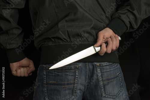 Fotografie, Tablou hides a big knife behind his back in young man hand of a black jacket and jeans clenched fist