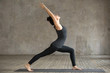 © fizkes - Young woman practicing yoga, doing Virabhadrasana 1 exercise, Warrior one pose, working out, wearing sportswear, black pants and top, indoor full length, gray wall in yoga studio