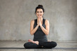 © fizkes - Young smiling woman practicing yoga, doing Padmasana exercise, Lotus pose, working out, wearing sportswear, black pants and top, indoor full length, yoga studio window, looking at the camera
