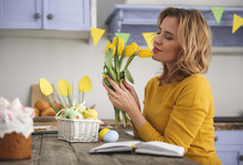 Profile Of Serene Attractive Woman In Kitchen Holding Yellow Tulips And Smiling With Eyes Closed. Decorated Straw Basket Filled With Colorful Eggs Is Standing On Table. Copy Space In Left Side