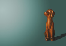 Wooden, Gold Doggy Figurine. The Wooden Dog Looks Up With A Satisfied Face. A View Of A Dog Who Sits On A Dark Pastel Background. Animal Concept, Minimalism Concept.