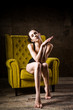 © Elizaveta - Young beautiful, sexy Caucasian woman with a thin figure and long bare legs, barefoot posing sitting on a yellow chair in the interior against a dark wooden wall. Dressed in black classic underwear