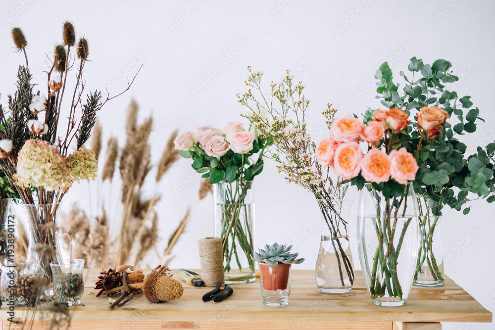 pink roses on a wooden table on which are located roses, dried flowers cones, greens, scissors, rope, bumps on a white wall background - obrazy, fototapety, plakaty 