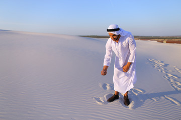 Wall Mural - Happy young Emirati Sheikh bends over and takes white sand in hands, rises up and lets sand through fingers of hands, smiling on face, standing in midst of bottomless desert with white sand on hot