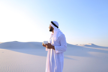 Portrait  confident and happy male Muslim who drinks invigorating coffee drink from white cup and enjoys calm morning, standing in midst of endless sandy desert with pure white sand in open air on war