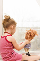 Little girl with doll looking out the window of the house